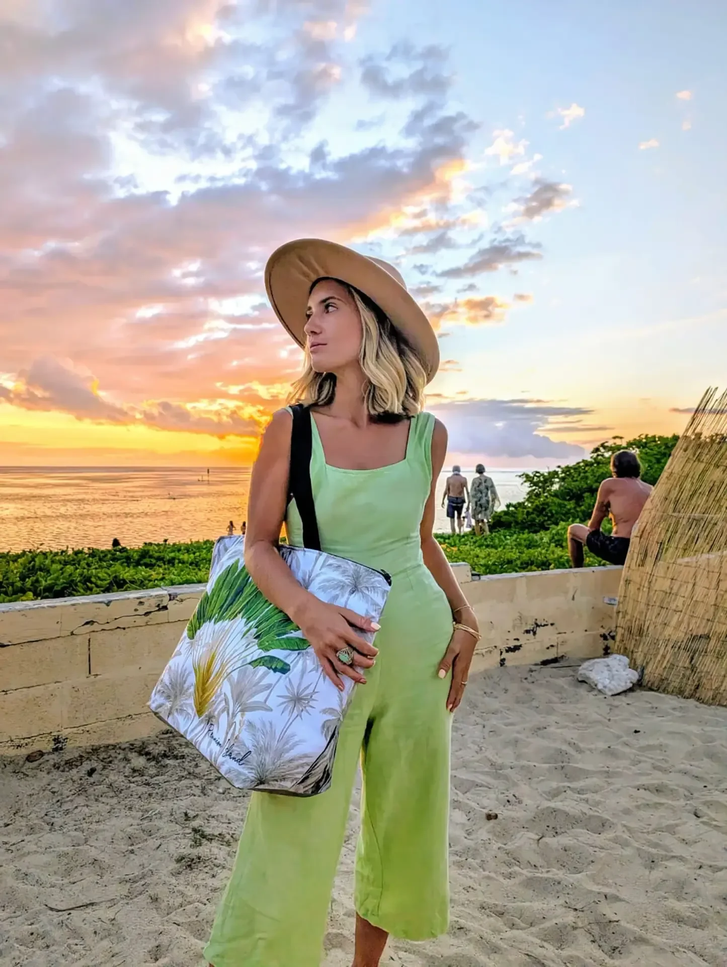 Au coucher du soleil sur une plage de sable, une femme en combinaison verte et chapeau porte à l'épaule un cabas Ravenala illustré de feuilles de l'arbre du voyageur