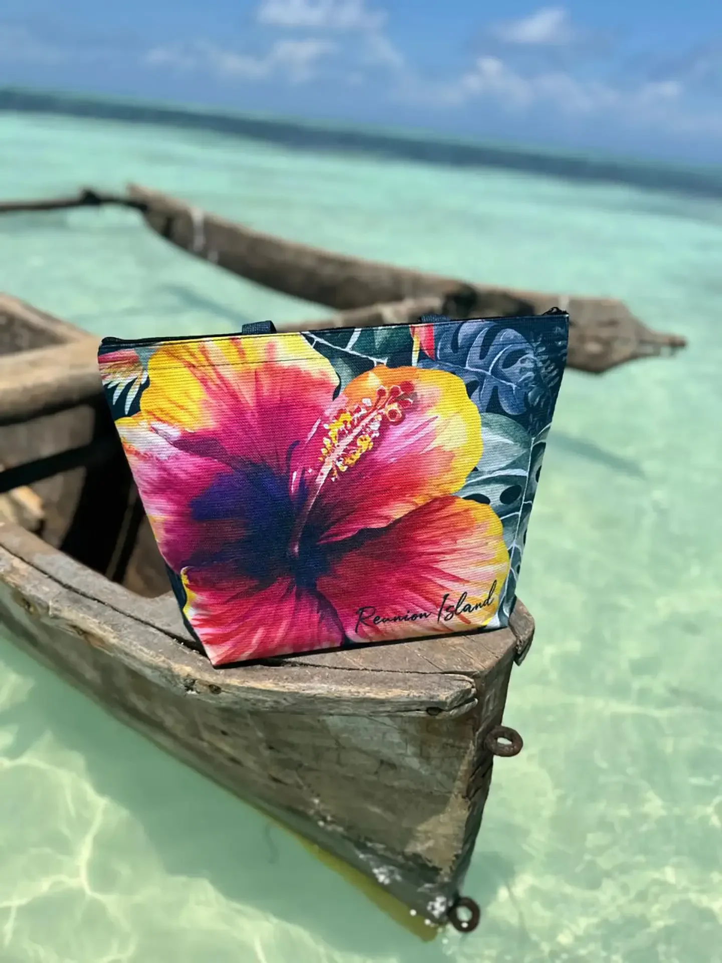 Un cabas Zanzibar illustré d'une grande fleur d'hibiscus rose et jaune posé à l'avant d'une pirogue en bois, sur une eau turquoise et transparente