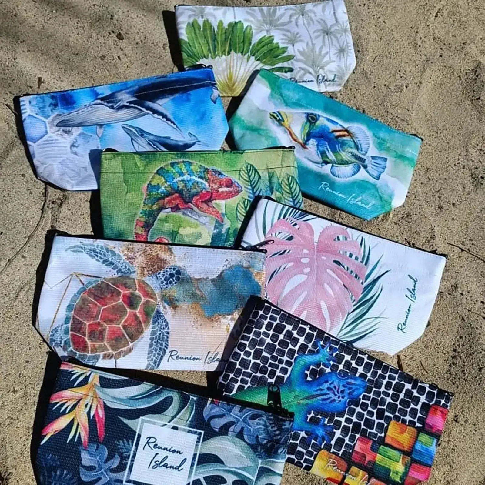 Plusieurs trousses Tropical Factory étalées sur le sable, aux motifs de baleine, caméléon, tortue marine, poisson baliste, feuilles de monstera et ravenala