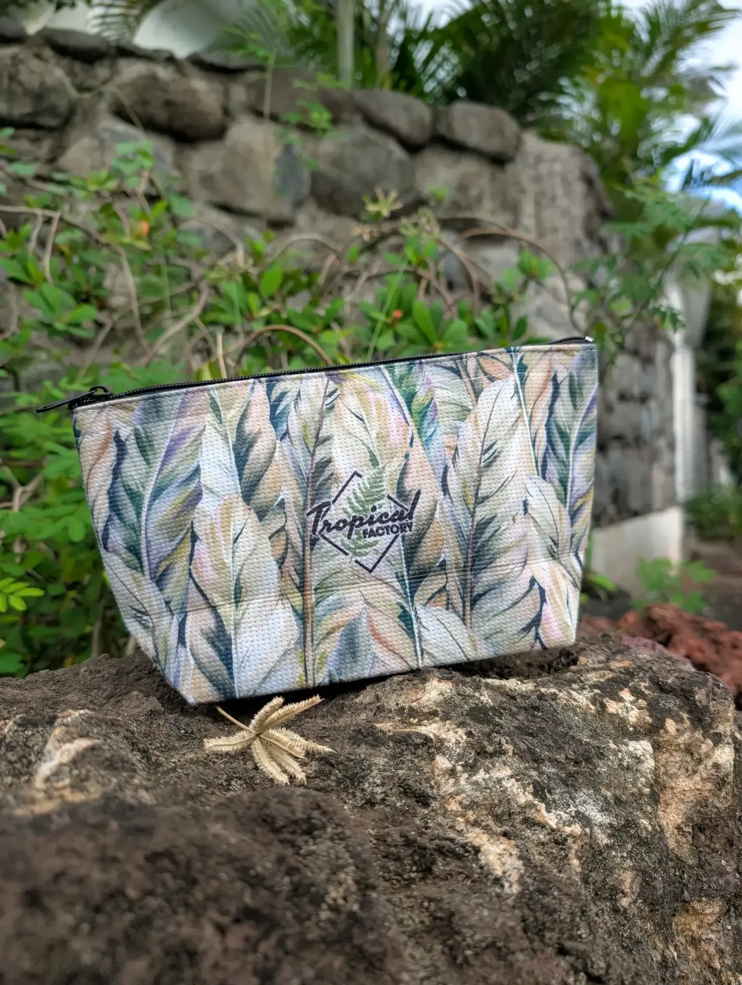 Une trousse Tropical Factory aux motifs de longues feuilles vertes, beiges et bleues, posée sur un rocher devant un mur de pierres et de la végétation
