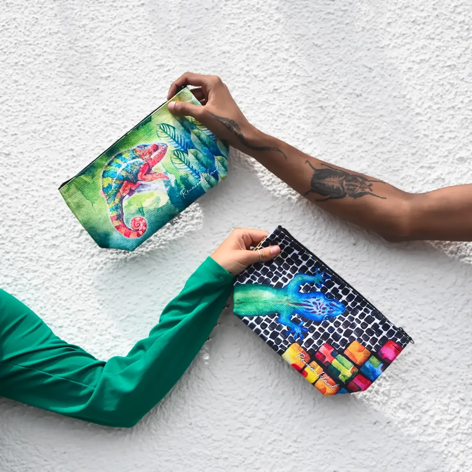 Deux bras tendus devant un mur blanc tiennent chacun une trousse Tropical Factory, l'une illustrée d'un caméléon coloré, l'autre d'un lézard bleu sur fond de mosaïque