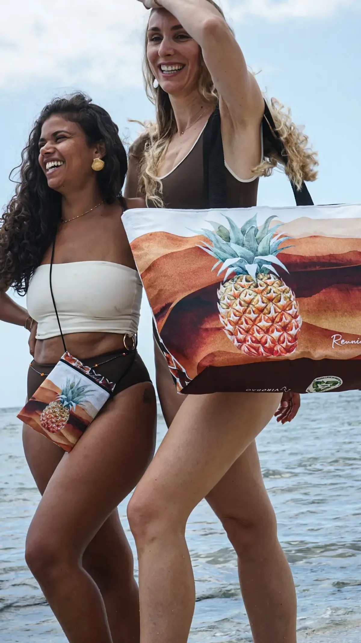 Deux femmes rieuses au bord de l'eau, l'une portant une pochette, l'autre un grand cabas Victoria illustré d'un ananas sur fond de dunes orangées