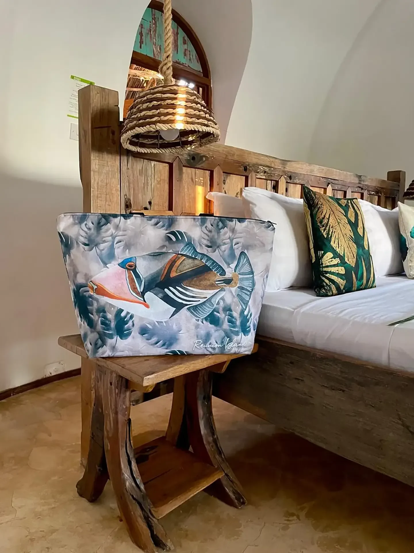Un cabas Tropical Factory illustré d'un poisson baliste posé sur un tabouret en bois, à côté d'un lit garni de coussins, sous une suspension en corde