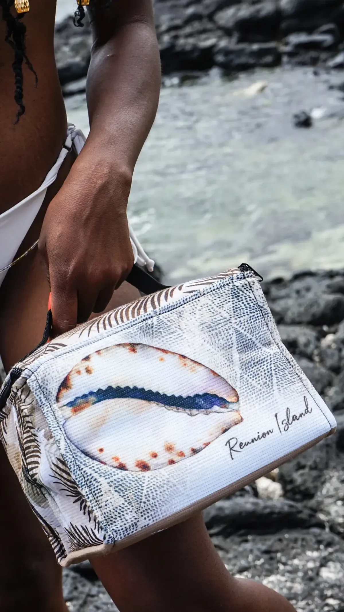 Une main tient par la poignée un petit sac Kori illustré d'un coquillage cauri et de feuilles grises, au-dessus de rochers noirs et d'une eau claire