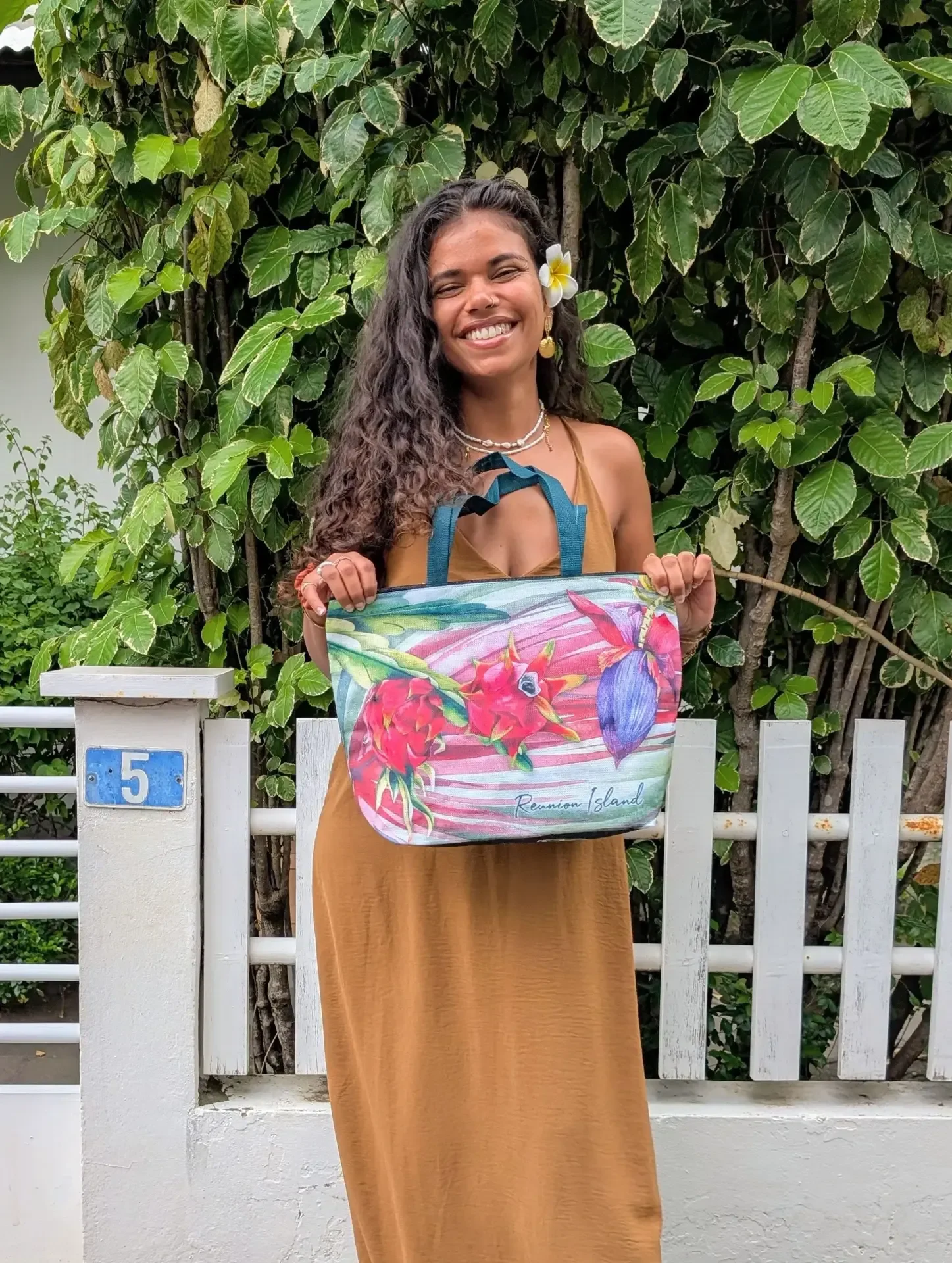 Une femme souriante, une fleur dans les cheveux, tient un cabas Pitaya aux fleurs roses et violettes devant un portail blanc portant le numéro 5