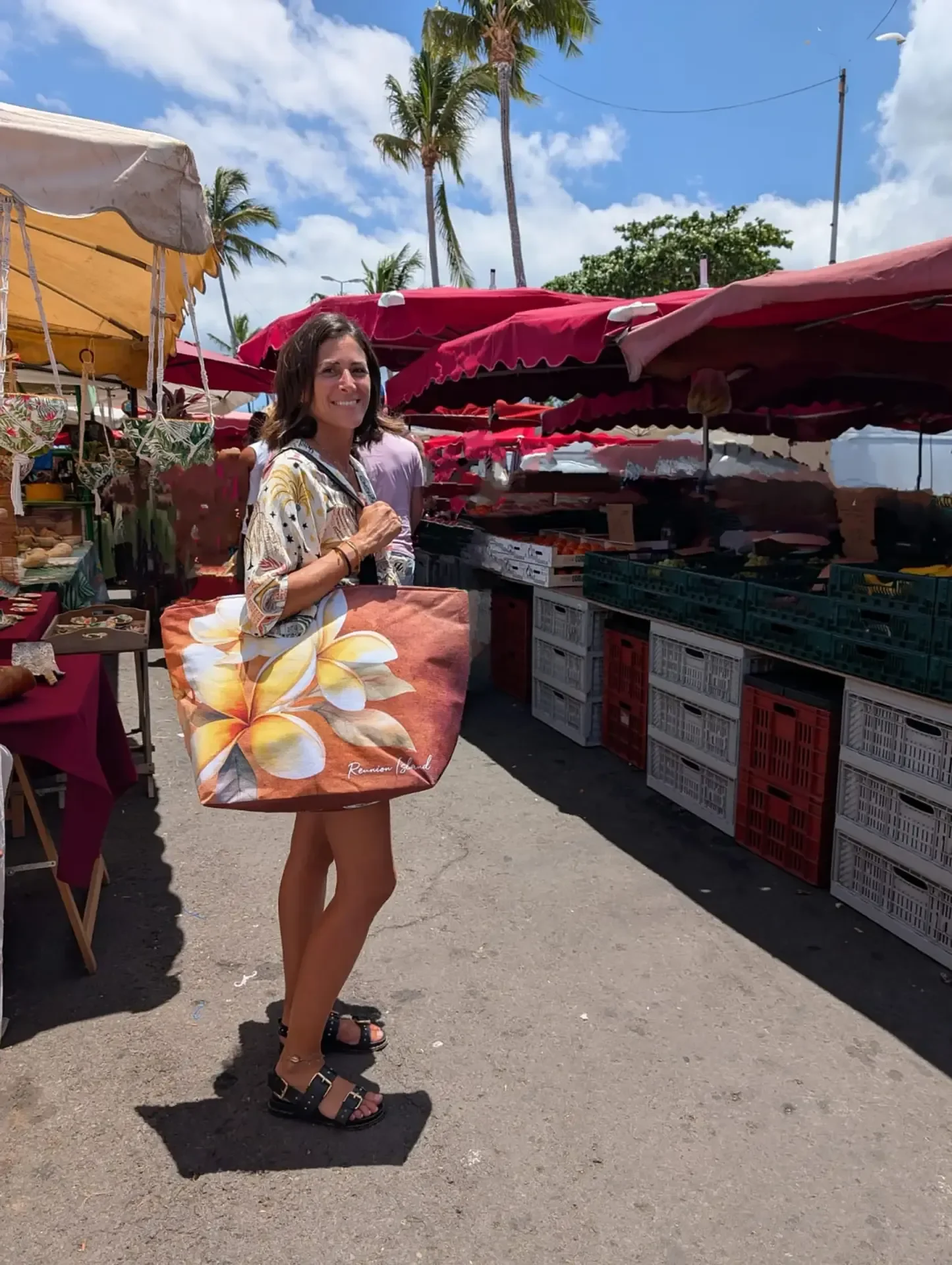 Une femme souriante porte un grand cabas Bora orné de fleurs de frangipanier dans une allée de marché, sous des parasols rouges et des palmiers
