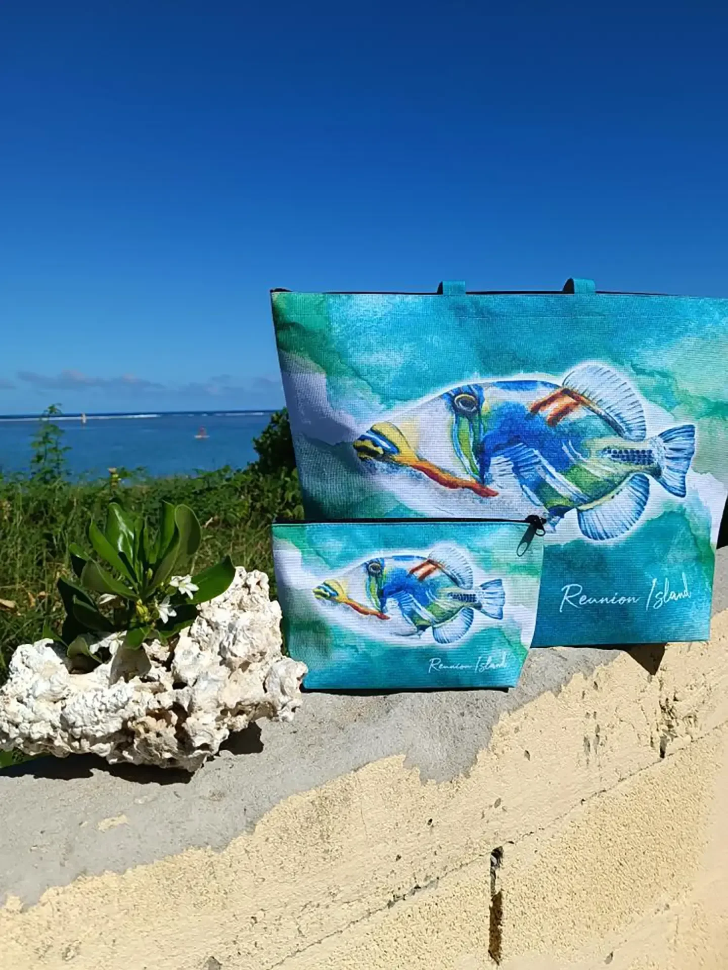 Un cabas et une trousse turquoise illustrés d'un poisson baliste Picasso posés sur un muret, à côté de morceaux de corail blanc, avec la mer au loin