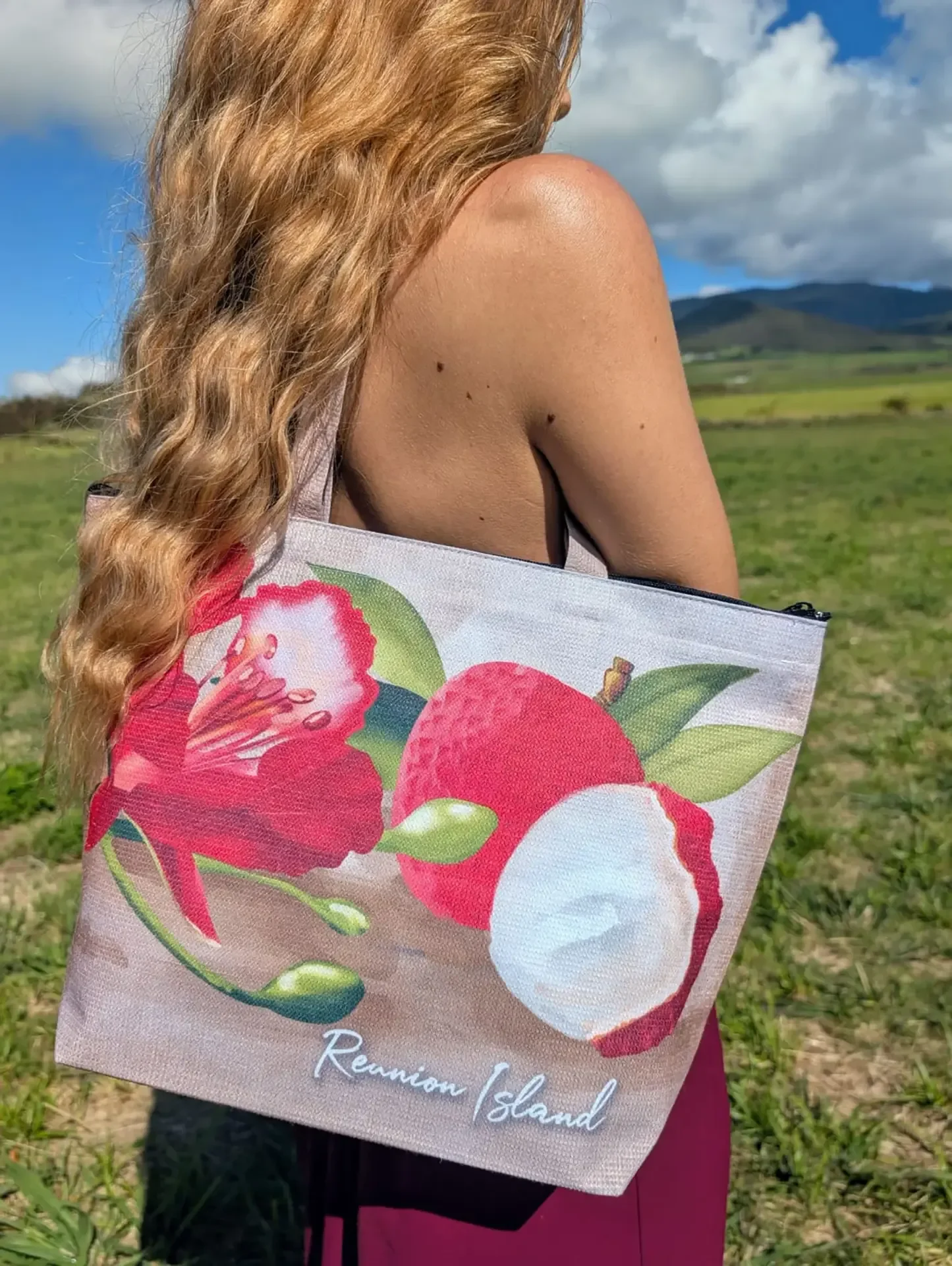 Une femme aux longs cheveux porte à l'épaule un cabas Letchi illustré de letchis et d'une fleur rouge, dans une prairie avec des montagnes au loin