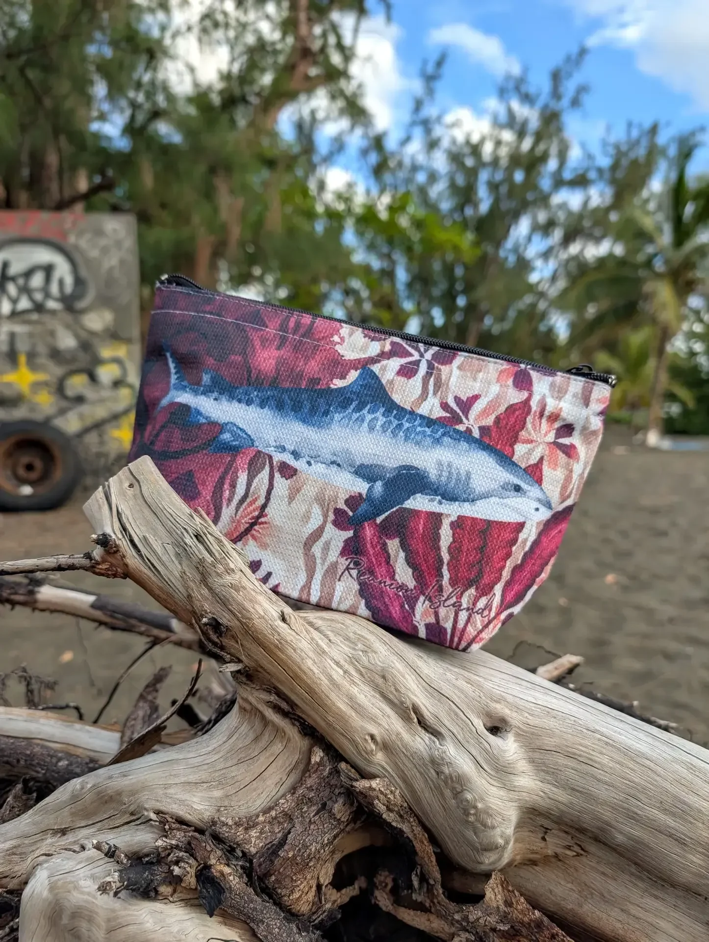 Un sac isotherme Nguzi illustré d'un requin tigre sur fond de fleurs rouges et roses, posé sur un tronc de bois flotté, sur une plage de sable noir