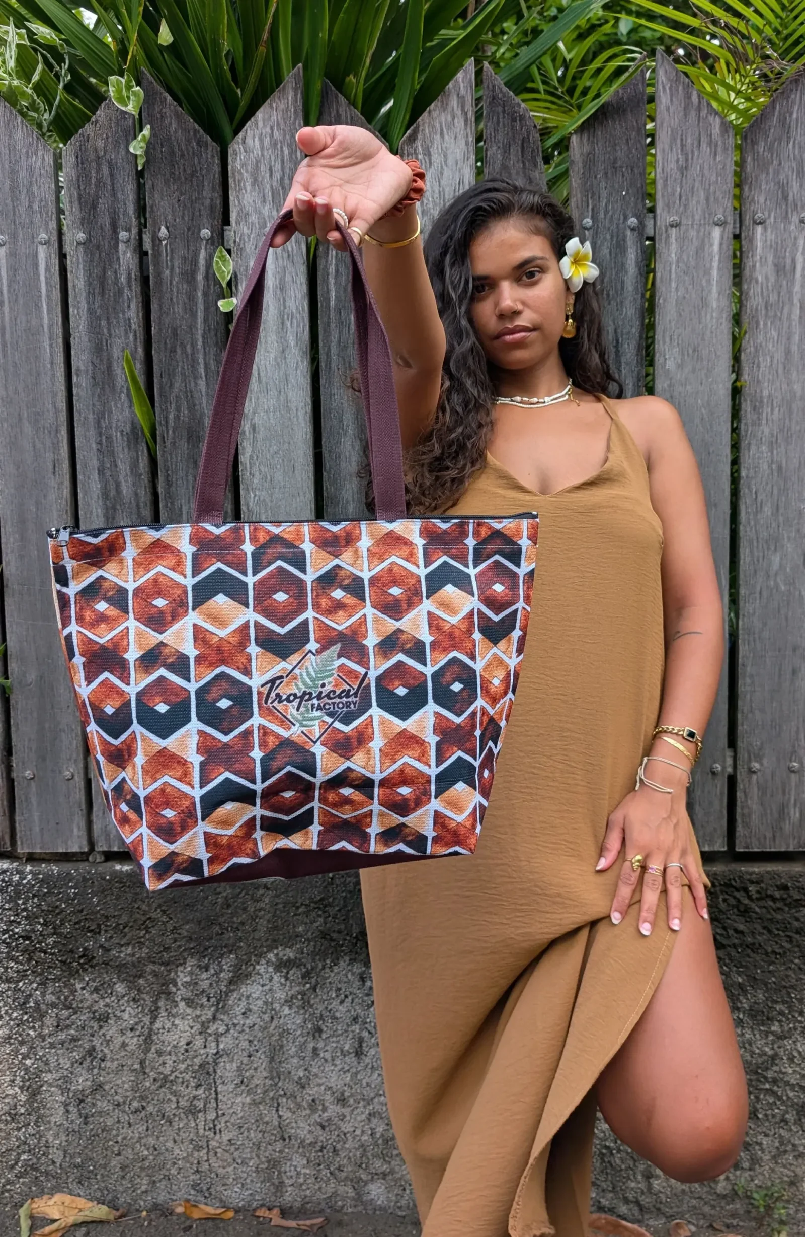Devant une palissade en bois, une femme en robe camel tend à bout de bras un cabas Tropical Factory à motif géométrique orange, brun et bleu nuit