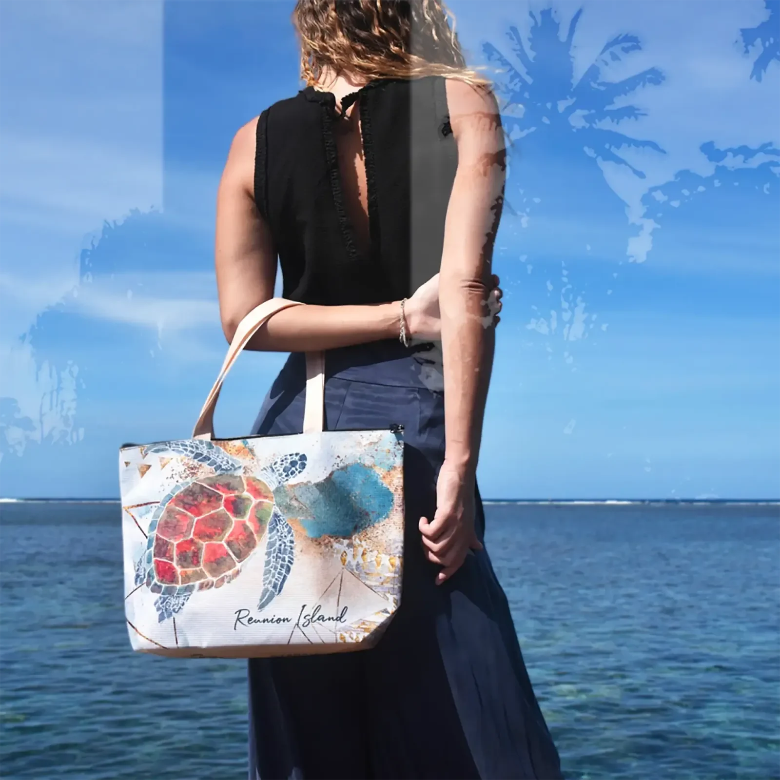 Vue de dos, une femme en haut noir tient un cabas Fano illustré d'une tortue marine aux tons rouges et turquoise, face à l'océan sous un ciel bleu