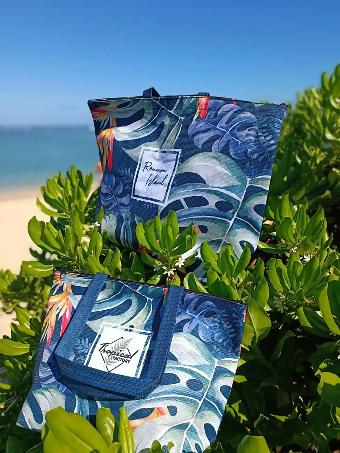 Deux sacs Tropical bleu nuit illustrés de grandes feuilles et de fleurs orangées, posés sur un buisson vert face à la mer